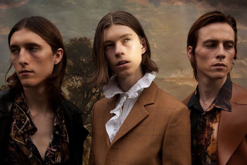 Three young men wearing shirts with extravagant collars pose in front of a landscape painting.