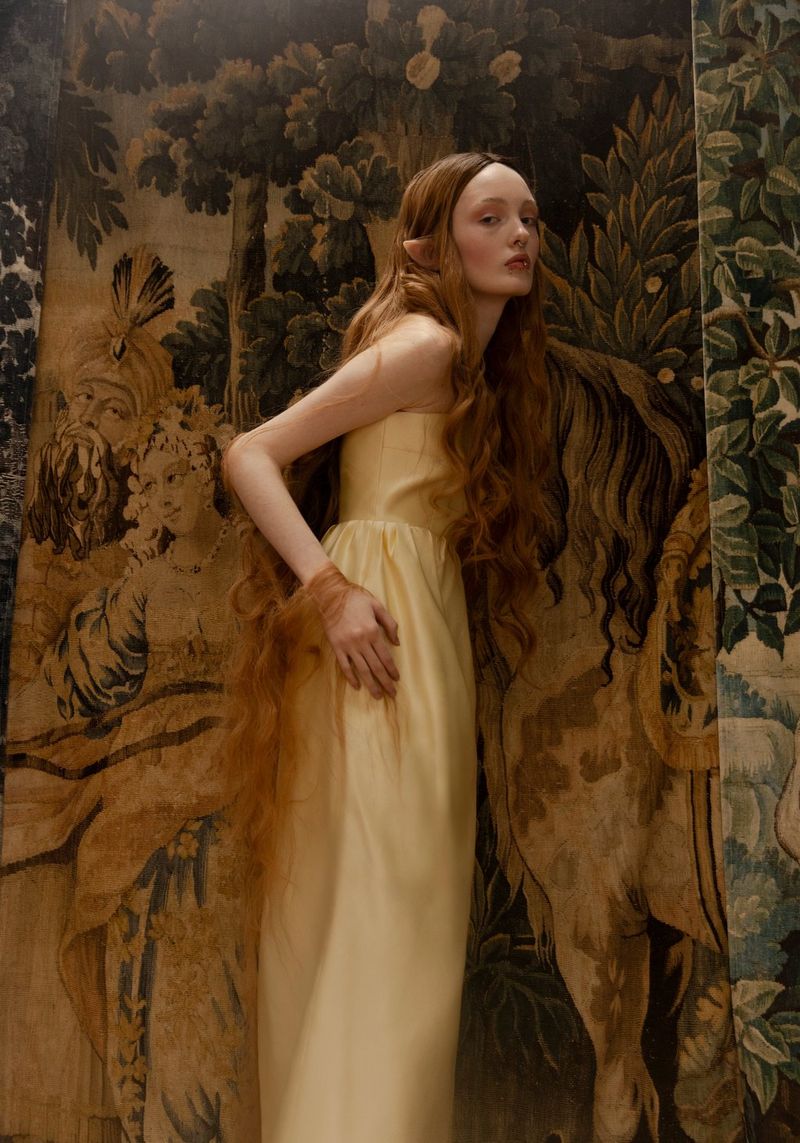 A fashion portrait of a woman with elven ears and very long red hair, wearing a long, pale yellow dress, standing in front of a tapestry.