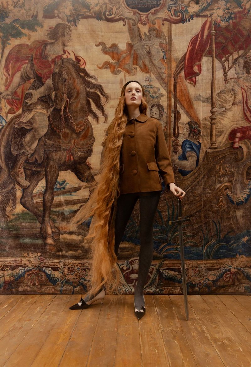 A fashion portrait of a woman with elven ears and very long red hair, wearing a brown jacket and holding a cane, standing in front of a tapestry.