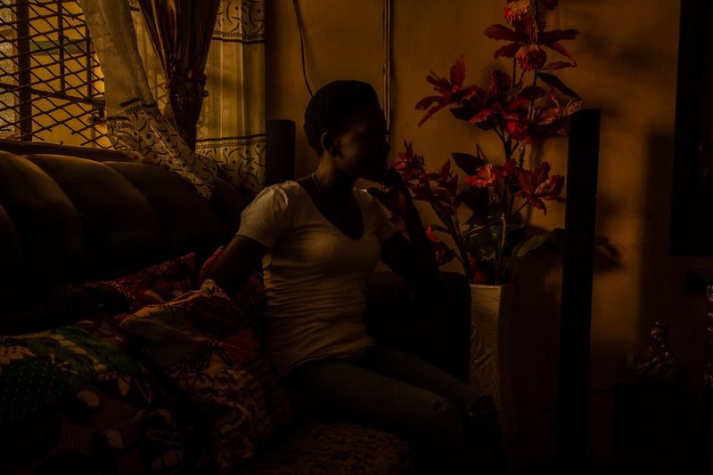 A teenage mum rests her chin on her hand while sitting on a sofa in a darkened room.