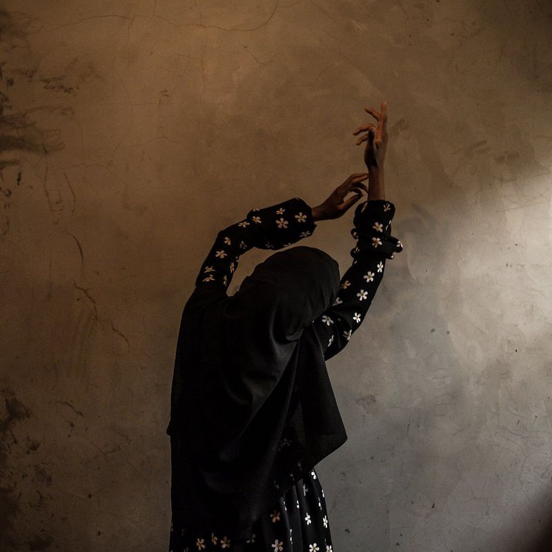 A woman in a headscarf stands against a wall with her back to the camera and her arms raised. 
