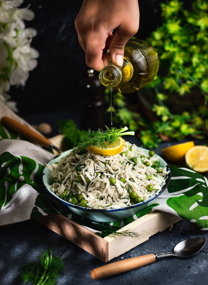 A hand pours oil over a dish of rice and peas topped with a lemon and garnished with herbs. 