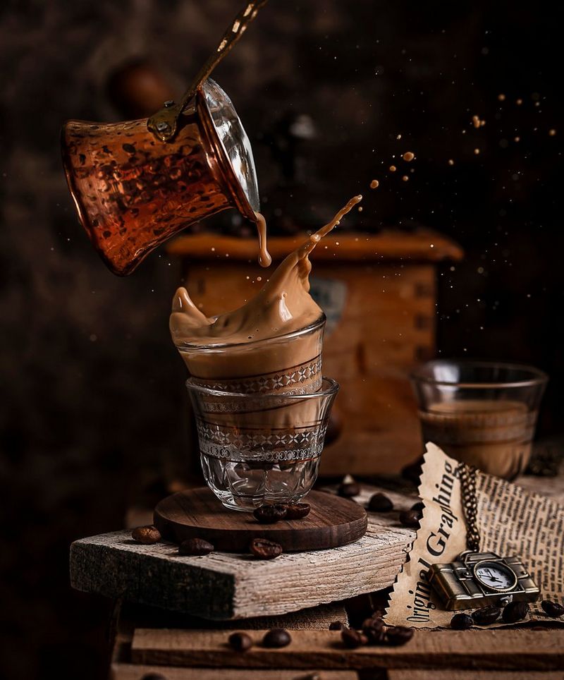 Coffee splashes over the rim of a glass as it's poured from a copper ladle.