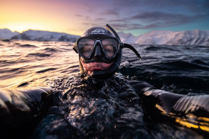 Canon Ambassador Stéphane Granzotto half-submerged in the sea, wearing a wetsuit and snorkel and holding out his hands to take a selfie with his Canon EOS R5 using underwater housing. 
