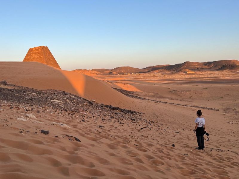Canon Ambassador Camilla Ferrari stands amid sand dunes in Sudan, a pyramid with a broken top nearby.
