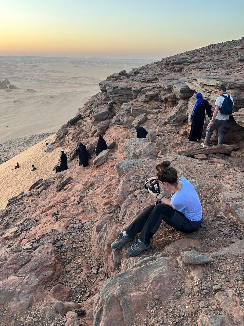 Canon Ambassador Camilla Ferrari sits on a rocky ledge in Sudan, using her Canon EOS R5 to take photographs of people descending the rock to the beach.