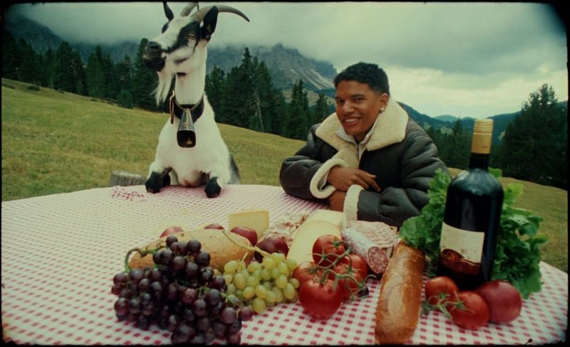 Рэпер AntsLive сидит за уличным столом, слева от него можно увидеть еду, справа — козу, закинувшую копыта на стол, а позади виднеется горный пейзаж; снято с помощью объектива Super-16 Canon SC15x11.