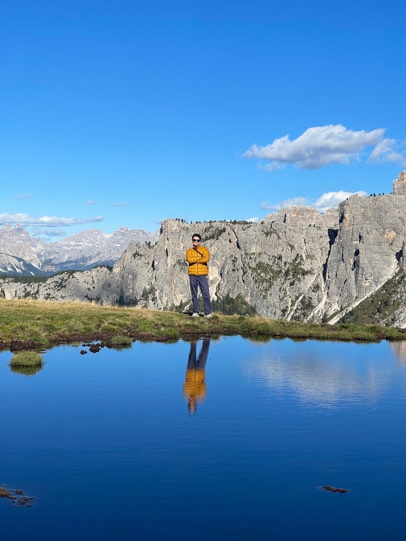 AntsLive стоит возле водоема на фоне Доломитовых Альп; в воде можно увидеть отражение музыканта и синего неба.