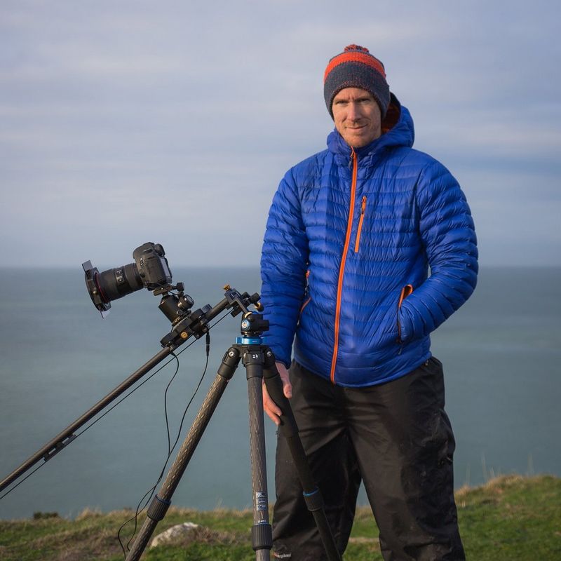 Landschapsfotograaf Drew Buckley in winterkleding; hij staat op een met gras bedekte rots en achter hem is de zee zichtbaar. Naast hem staat een statief waarop een Canon-camera is bevestigd.