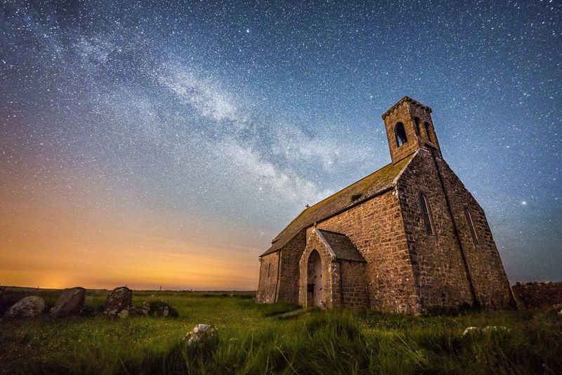 Een nachtelijke hemel gevuld met fonkelende sterren boven een kapel en een oud kerkhof.