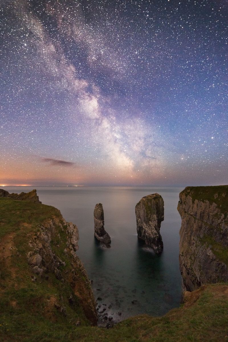 De hemel bij schemering, met de Melkweg boven hoge rotsen die uit de zee oprijzen.