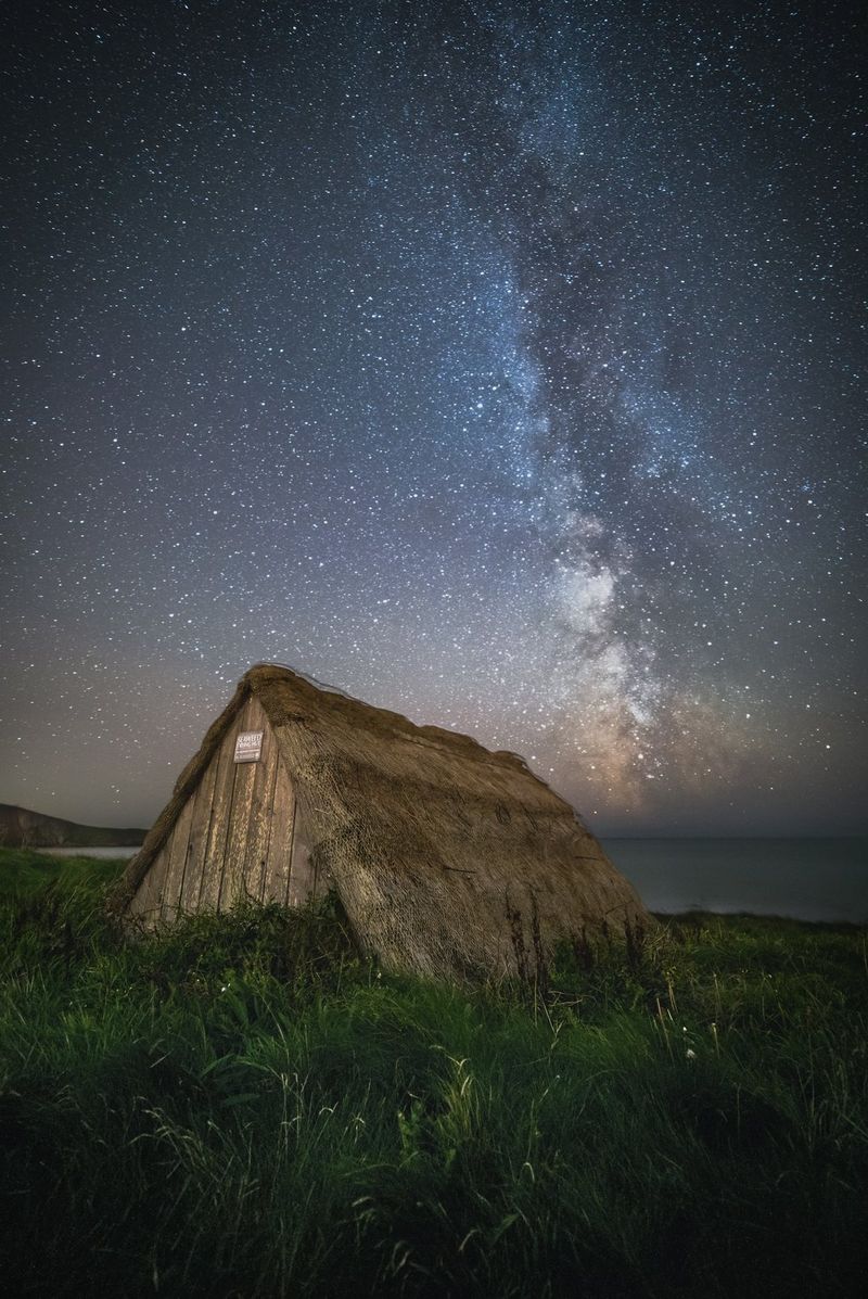 Een nachtelijke hemel met de Melkweg hoog boven een hut met een rieten dak aan de kust van Wales.