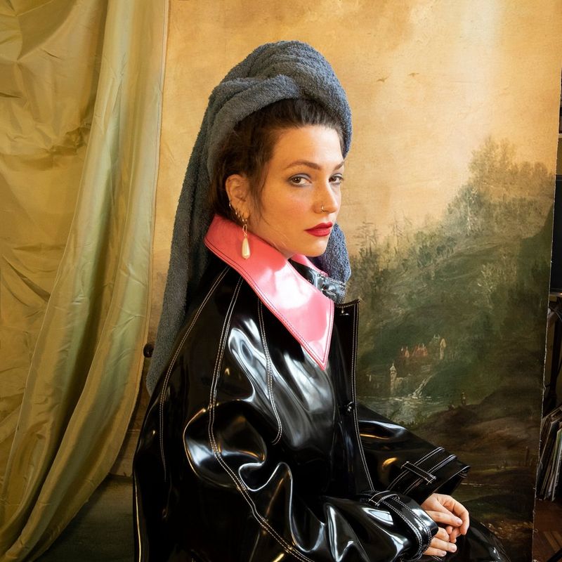 Un autoritratto della fotografa di moda e ritratti Wanda Martin, che indossa una giacca nera con un colletto rosa, un lungo orecchino di perle e un asciugamano avvolto intorno alla testa.