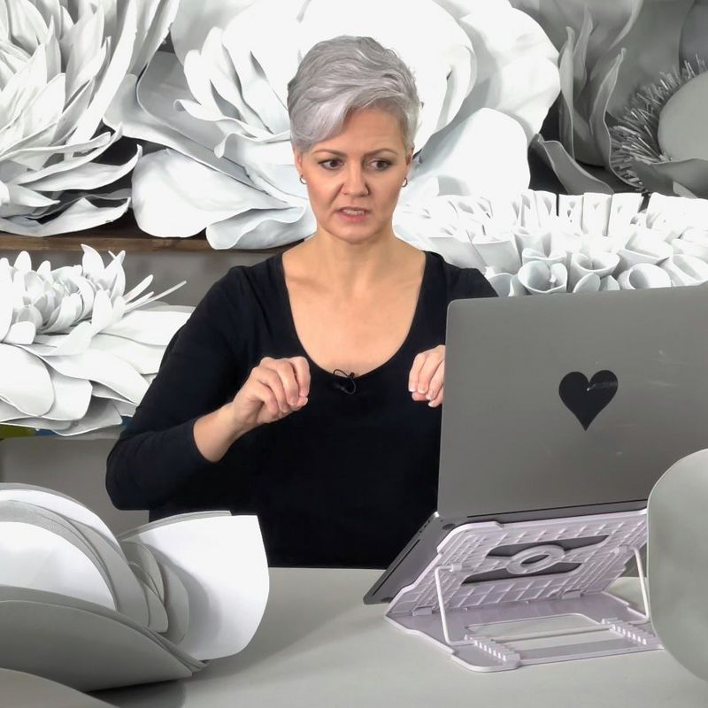  Newborn photographer Kelly Brown looks at her laptop inquisitively while seated at a small desk. Surrounding her are large, white, artificial flowers.