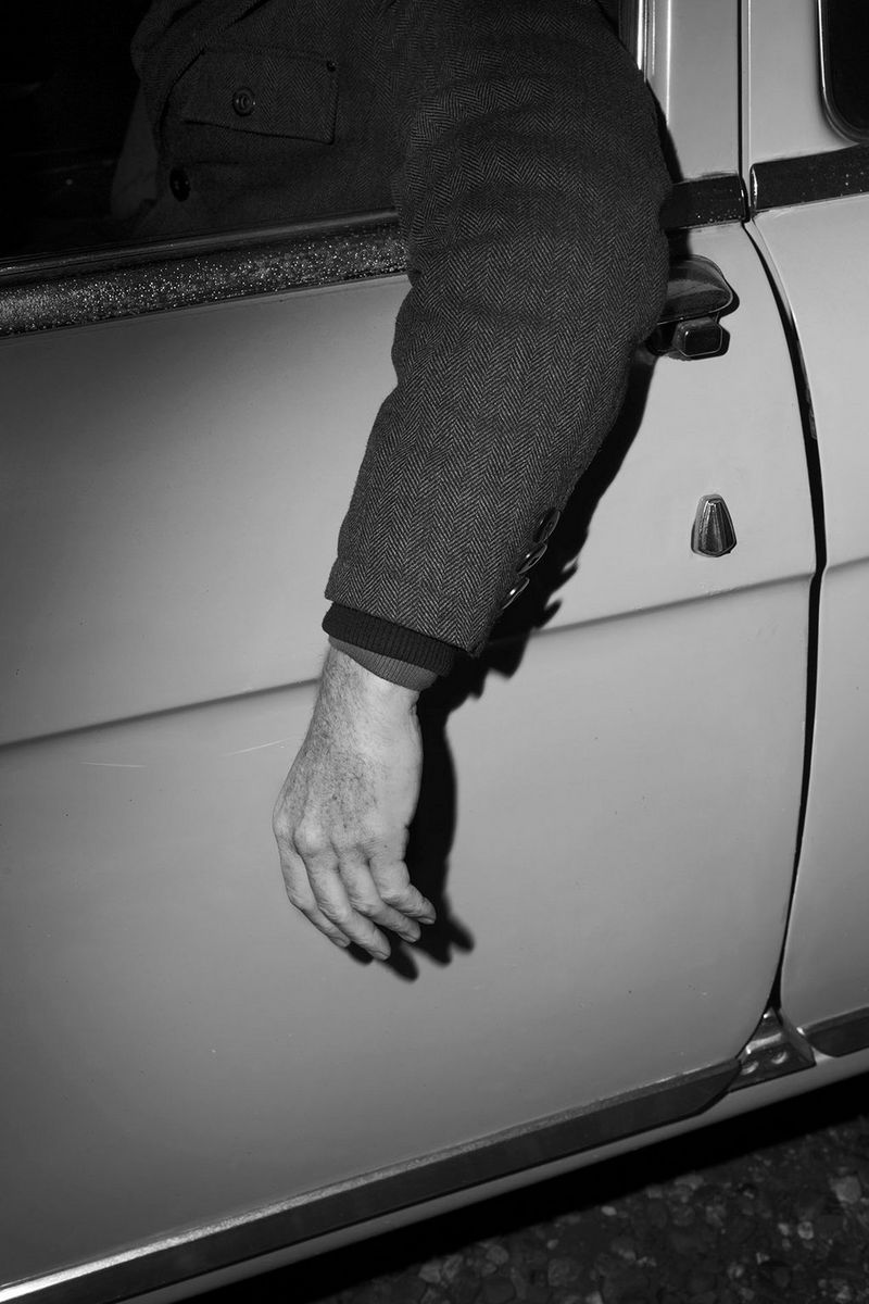 An arm in a herringbone jacket dangles out of the window of a car door, shot in black and white.