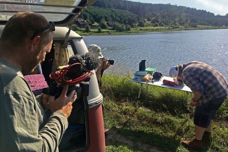 Clemens Boecker stands by the open tailgate of a VW campervan filming Joey Kelly cooking on a trestle table by a lakeside.