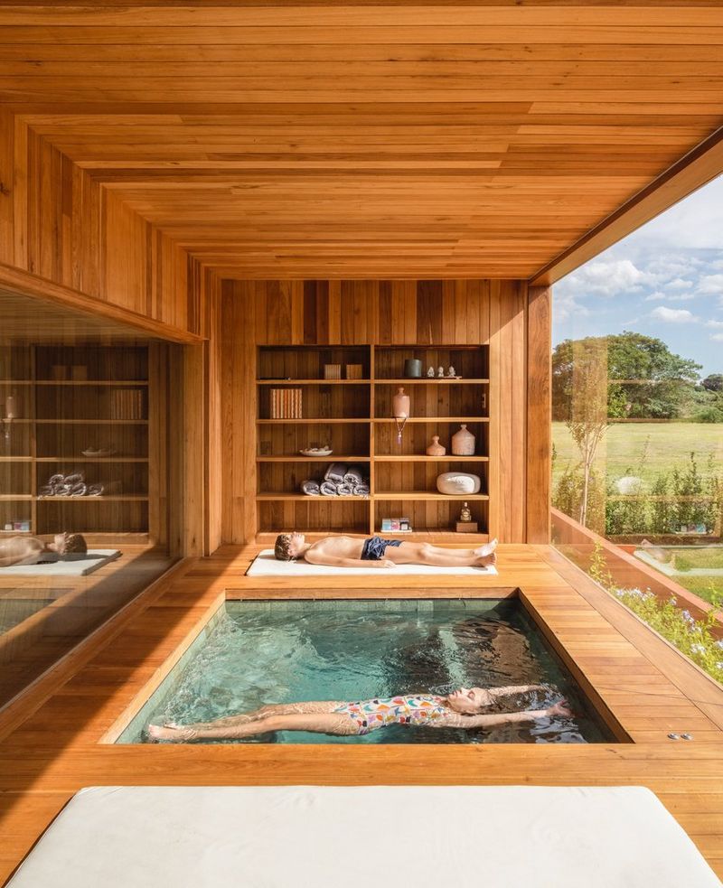 Two children relax in an elegant room with an indoor pool set into the wooden floors and a floor-to-ceiling glass window overlooking beautiful countryside. 
