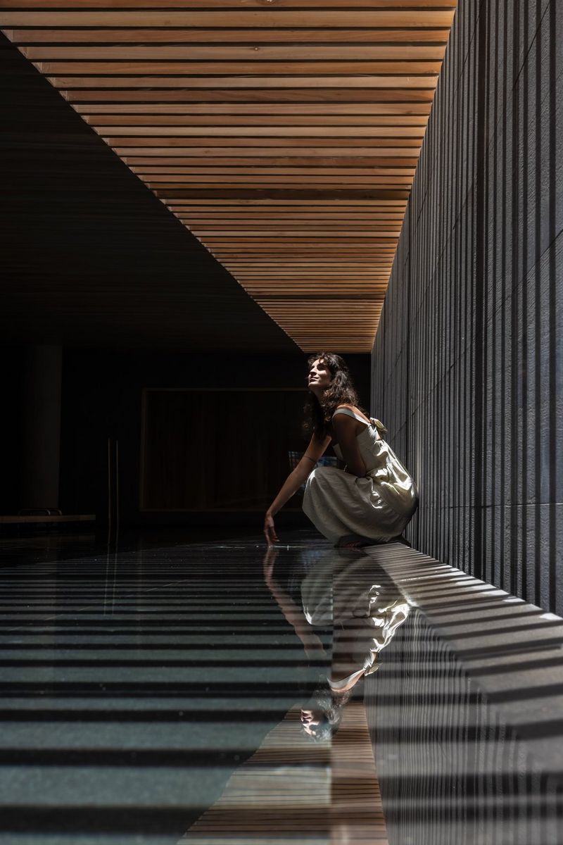 A figure crouches beneath a slatted wooden roof, which casts lines of shadow across the wall and floor.