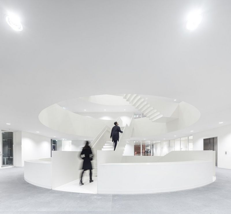 Two figures ascend a circular stairway in the centre of a large white room.