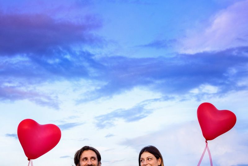 Una coppia tiene in mano dei palloncini rossi a forma di cuore contro un cielo blu/viola acceso. L'immagine è stata scattata in modo tale che si veda solo la parte superiore del volto dei soggetti.