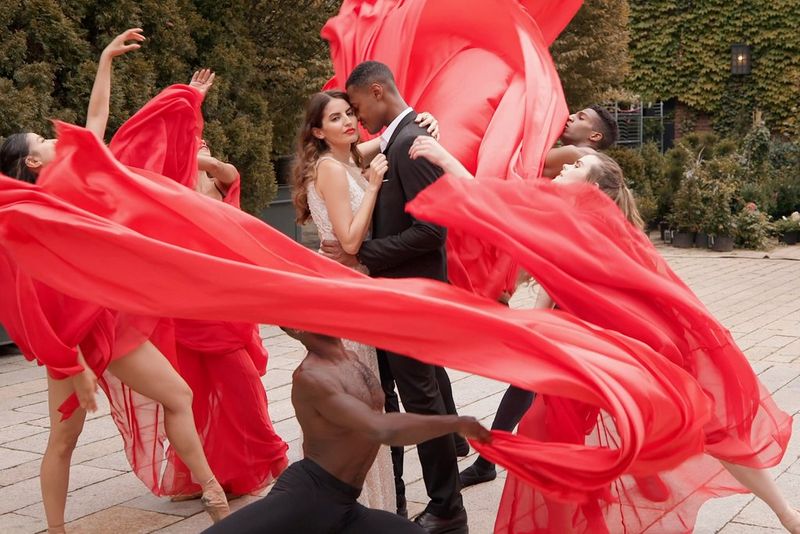 Una coppia si abbraccia in un giardino mentre un gruppo di ballerini le agita intorno dei drappi rossi.