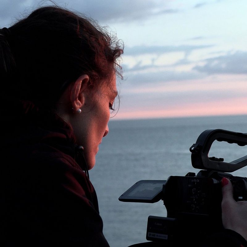 Un primo piano della regista Elisa Iannacone mentre guarda il touch screen orientabile di una videocamera Canon EOS C70 con davanti a sé una distesa d'acqua e un orizzonte dalle sfumature rosa.
