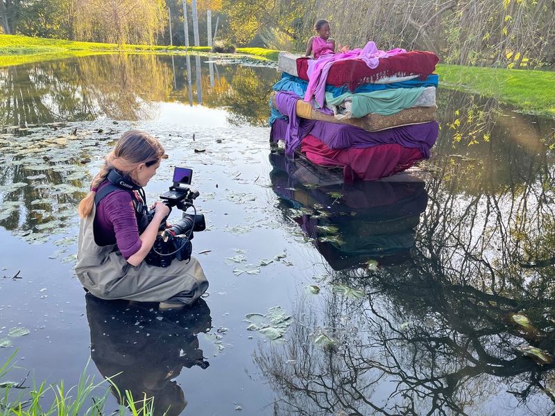 Filmmaker Elisa Iannacone crouches down in shallow water as she uses a Canon EOS C300 Mark II camera to film a young person sitting on top of a colourful pile of mattresses. 