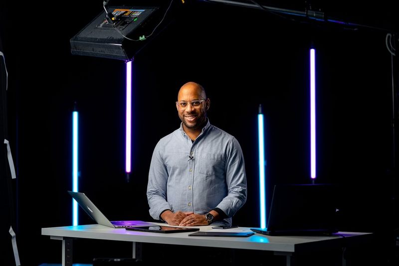 A man stands at a desk in a dark studio framed by four vertical strip lights.