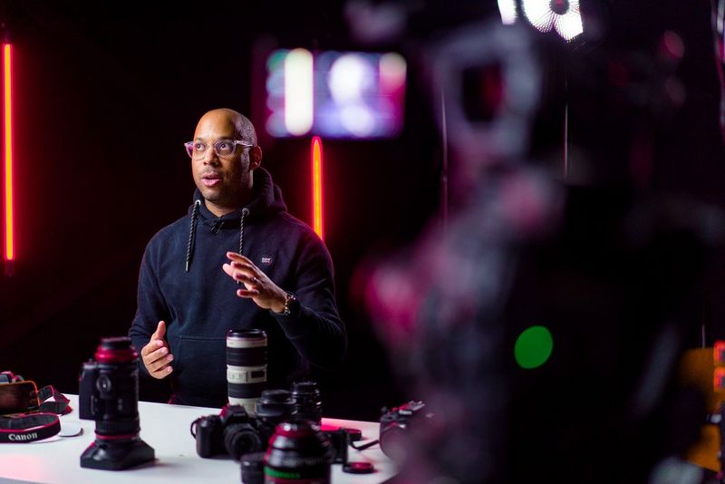 A man sits at a desk with several large Canon telephoto lenses plus some Canon cameras in front of him.