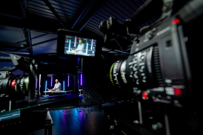 Две видеокамеры снимают мужчину, стоящего возле стола, — изображение дублируется на экране одной из камер.