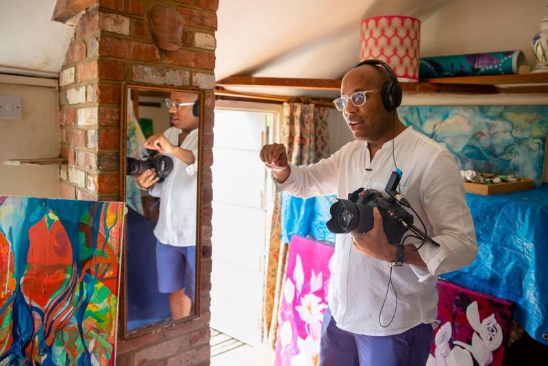 A man holds a Canon video camera to his chest, surrounded by paintings and reflected in a full-length mirror to his side.