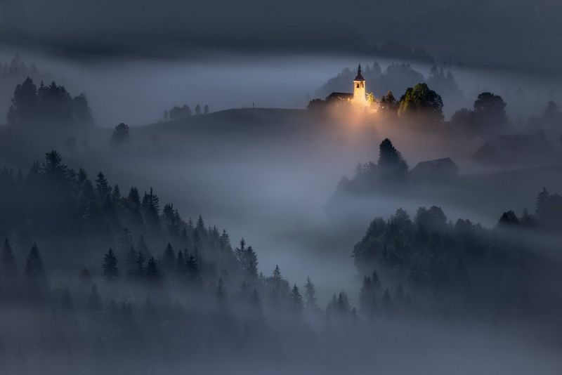 A misty nighttime landscape of tree-covered mountains, with a single lit-up church building in the distance, taken on a Canon EOS R5.