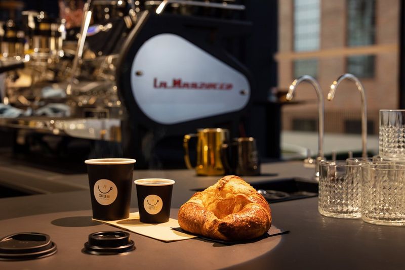 A croissant and two cups of takeaway coffee arranged on a cafe counter. A coffee machine can be seen in the background.