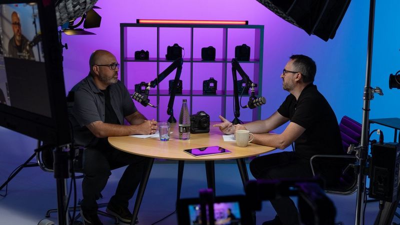 Фотограф спорта Ричард Хиткот и Майк Бернхилл из Canon сидят за круглым столом в студии на фоне витрины с камерами Canon.