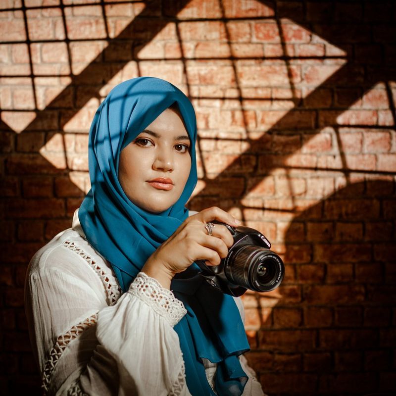 Yasmin Albatoul, in een witte blouse en blauwgroene hoofddoek, houdt een Canon EOS R-camera vast.