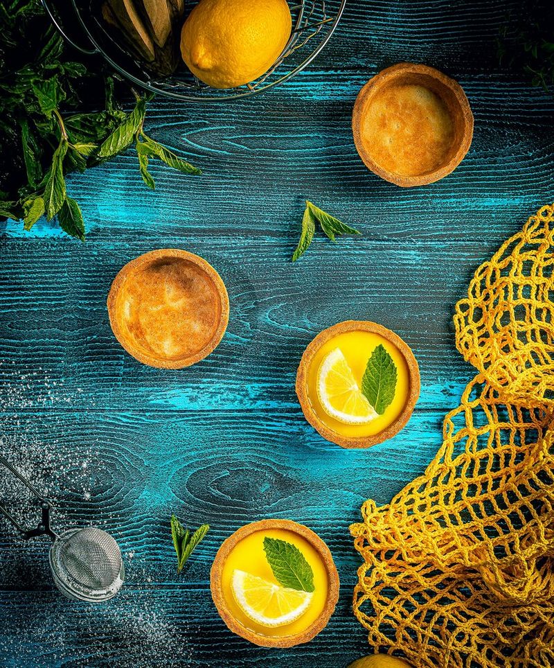 Een foto van Yasmin Albatoul van gebakken lekkernijen in het geel en oranje, bedekt met schijfjes citroen en muntblaadjes, op een helderblauwe, houten tafel.