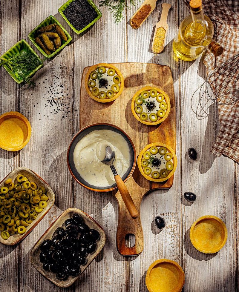 Een opname van bovenaf van Yasmin Albatoul van een tafel en snijplank vol ingrediënten en etenswaar, waaronder gehakte olijven, augurken en peperkorrels.