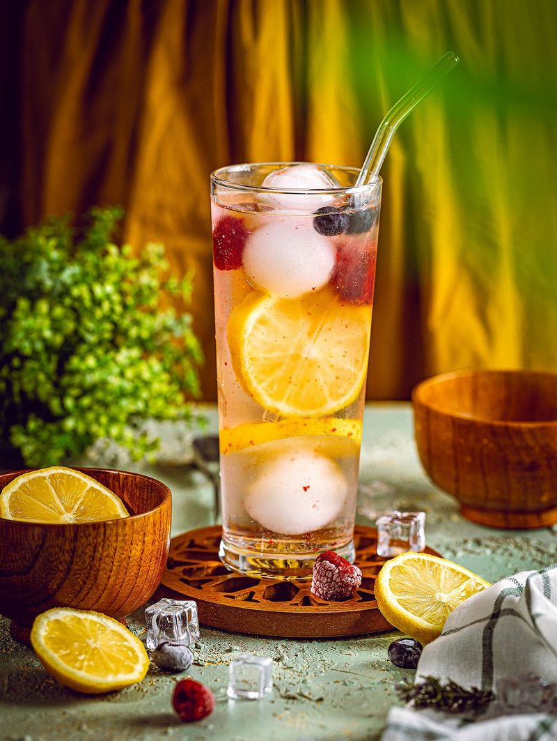 Een hoog glas met een vloeistof, ijs, fruit en een glazen rietje staat op een houten onderzetter, met daar omheen fruit en houten kommen.