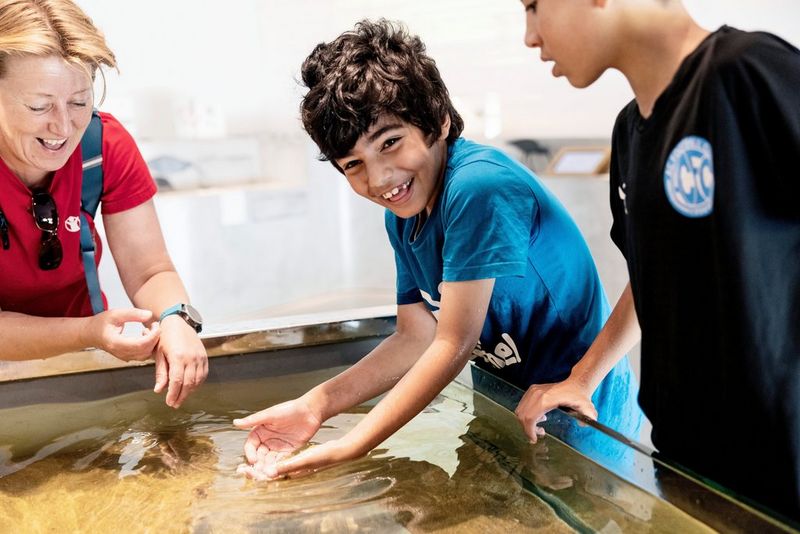 Een foto van Nanna Navntoft met twee jongens en een vrouw staande naast een watertafel. De jongen in het midden dipt beide handen in het water en kijkt lachend naar de camera.