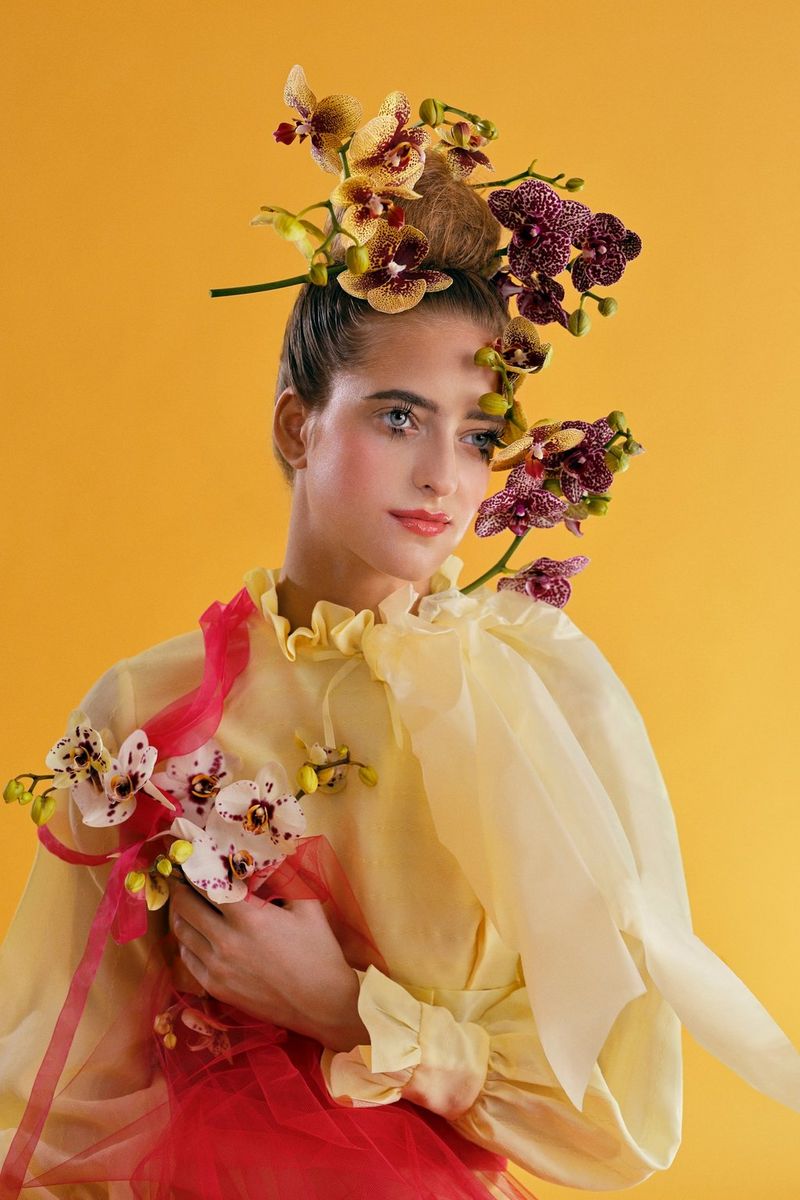 A model in front of a yellow background wearing large flowers on her head and neck.
