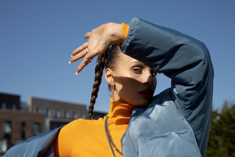 Модель в оранжевой водолазке и синей куртке держит руку в воздухе, создавая тень на своем лице.