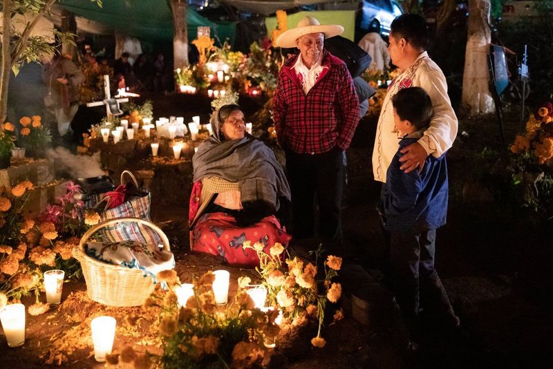 A group of people sit and stand at the edge of a square of earth arrayed with flowers, glasses with lit candles and a basket covered with a cloth. Behind them are other plots of earth, one with a cross at the edge of it.
