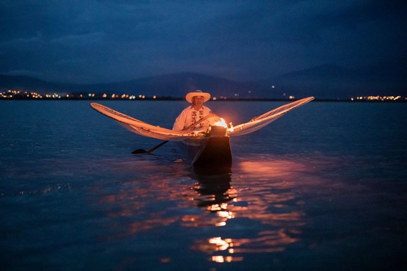 A man sits in a narrow boat with a large butterfly net stretching out either side of him. It is nighttime, and the boat is lit by a small fire at the front.