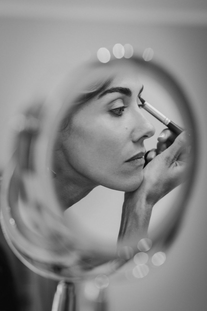 A black and white portrait of a bride in side profile, reflected in a mirror, applying make-up to her eyebrows.