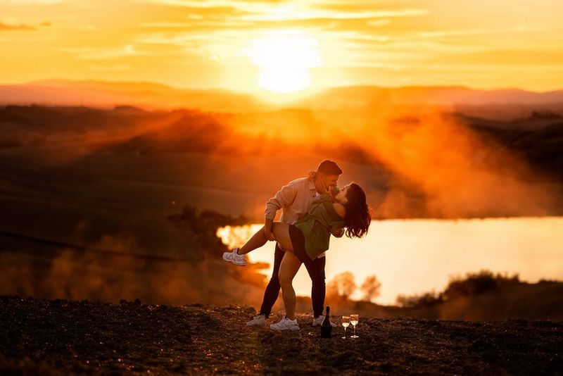 A couple embrace on a hillside as the sun sets behind them, a bottle of champagne and two glasses at their feet.