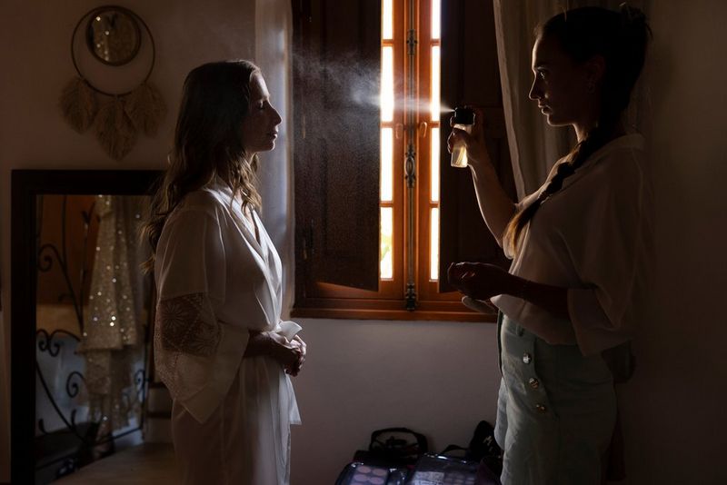 A make-up artist and a bride shot from the side in front of a shuttered window. The make-up artist is spraying a liquid onto the bride's face and the droplets are captured in mid-air by the light from the window.  