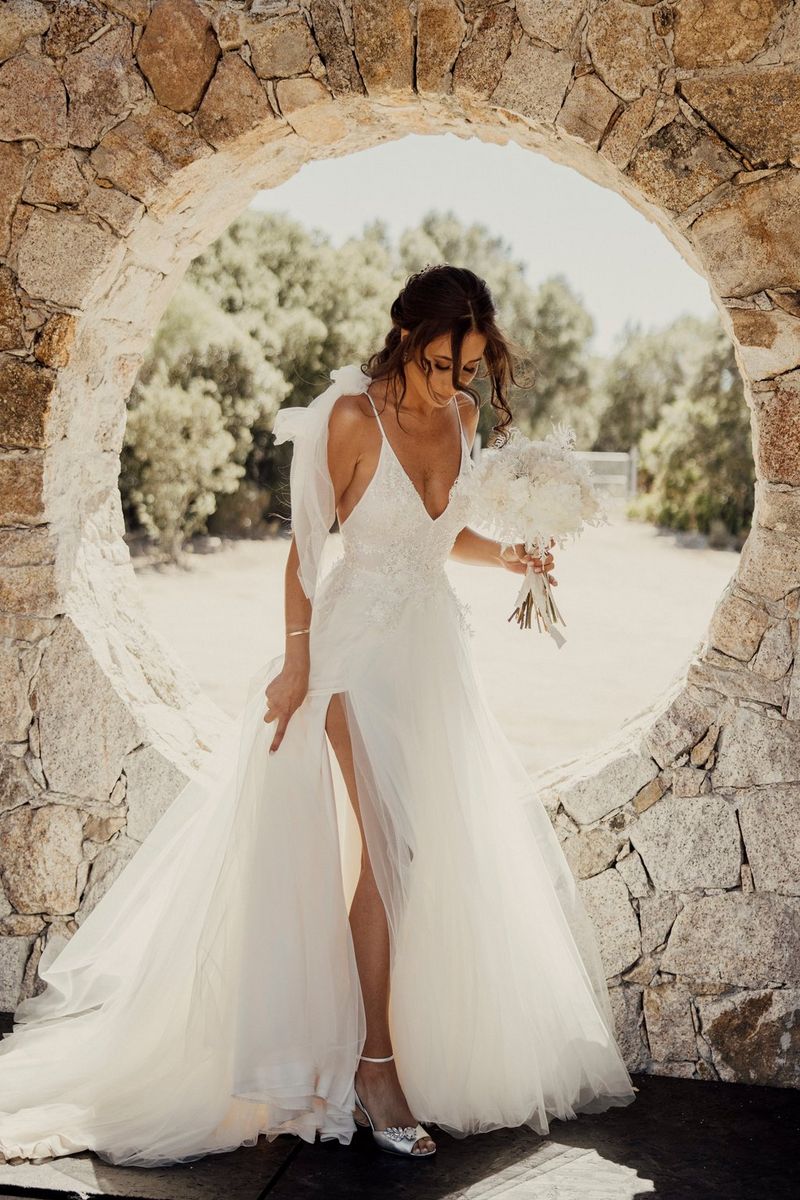 A bride in a long, flowing net dress, holding a bouquet of white flowers and looking towards the floor. She is standing in front of a circular stone window.