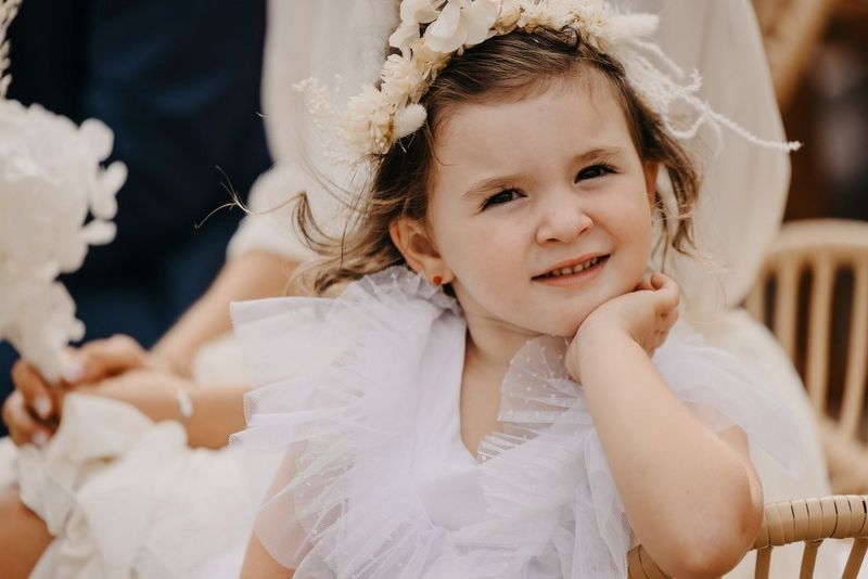 A portrait of a smiling flower girl in a ruffled dress leaning on the back of her chair and resting her chin on her hand.