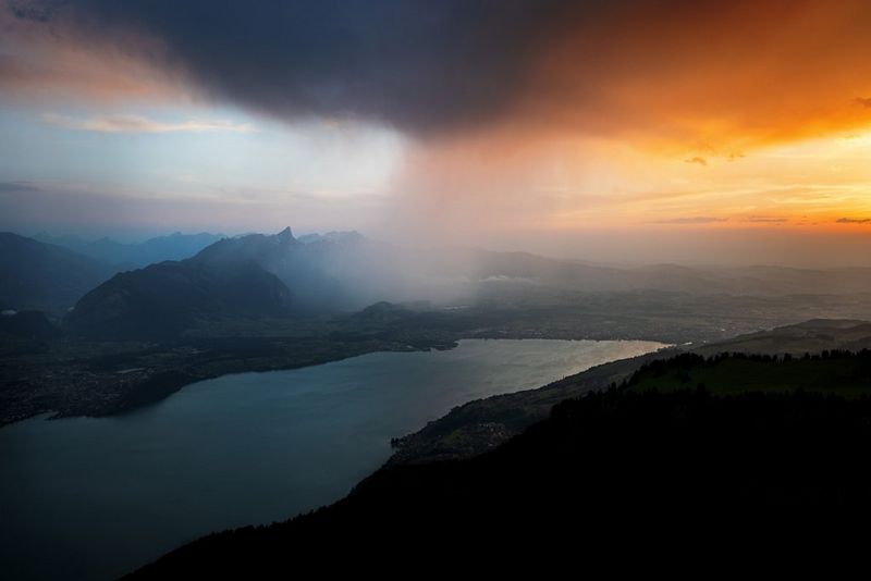Een zware regenbui trekt over een bergachtig gebied. Aan de ene kant van de stortbui is de lucht hemelsblauw, aan de andere kant goud-oranje.