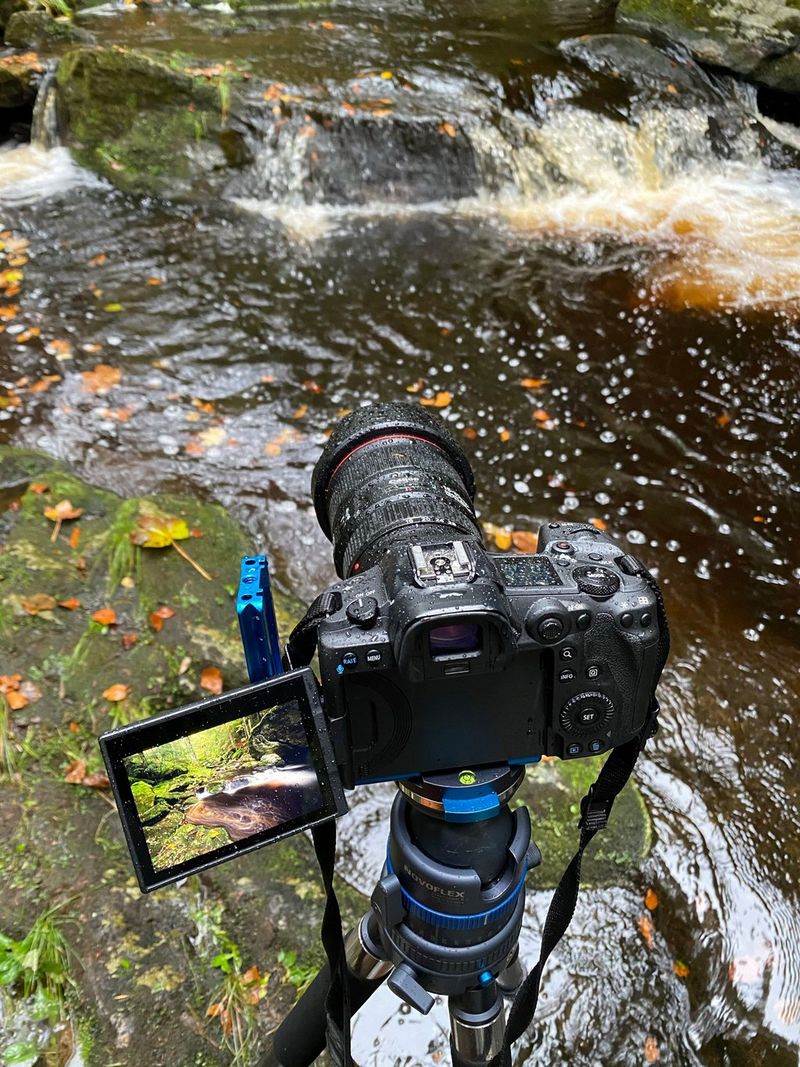 Een Canon EOS R5-camera op een statief in de regen, nabij een riviertje.